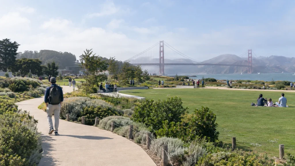Traveler walking through landscaped Presidio parkland with the Golden Gate Bridge visible in the distance.